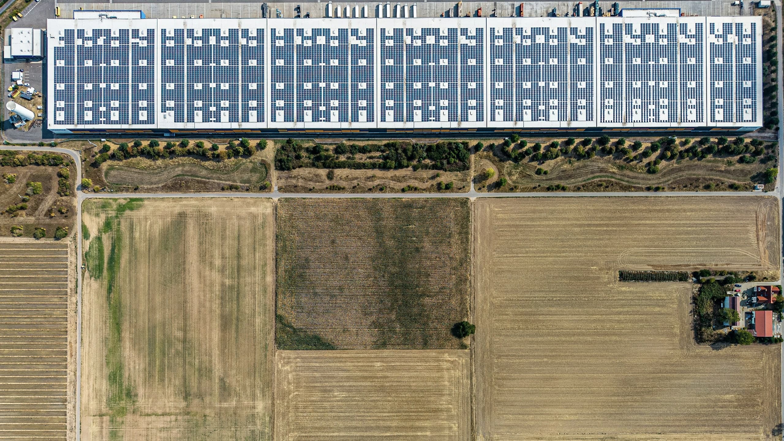 Top-down aerial view of a distribution warehouse whose entire roof is covered in solar panels, standing above flat, dry, harvested farmland.